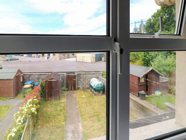 A view through a window of a residential area with a paved driveway, a small garden with flowers, and a green water tank. - 23 Cois Caol (Skibbereen)