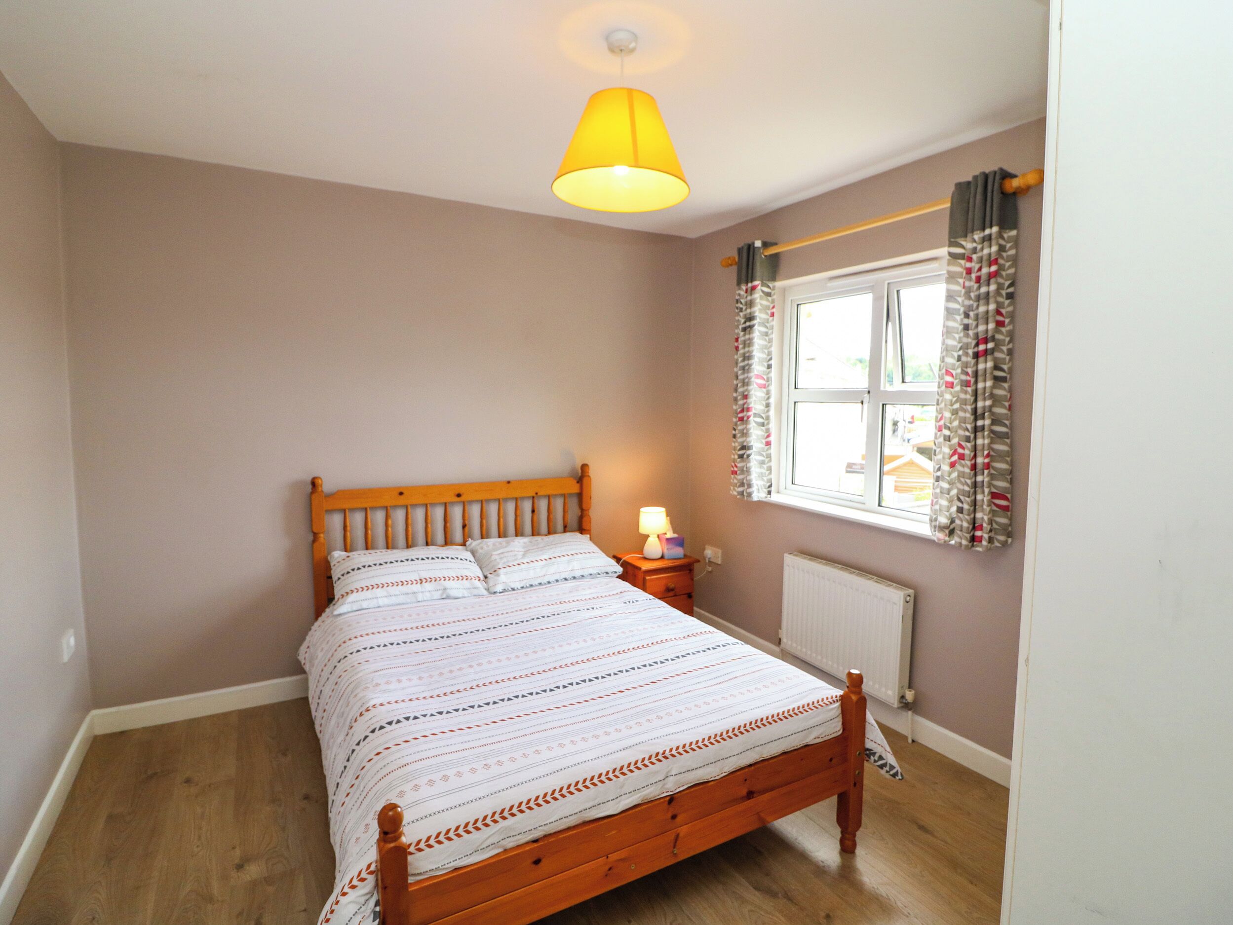 A bedroom with a wooden bed, a bedside table with a lamp, a window with curtains, and a radiator.