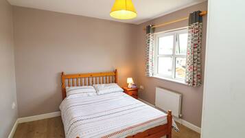 A bedroom with a wooden bed, a bedside table with a lamp, a window with curtains, and a radiator.