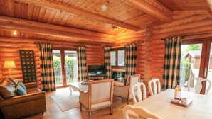 Interior de una cabaña de troncos con una mesa de comedor de madera, sillas blancas, un sofá y un televisor.