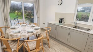 A dining area with a round table set for four, wooden chairs, a kitchen with a sink and kettle, and a view of the garden through a sliding door. - Folly Cottage (Narberth)