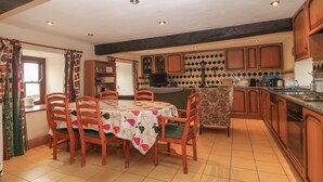 A kitchen with wooden cabinets, a tiled floor, and a dining area with a table and chairs. - Ereiniog 2 (Garndolbenmaen)