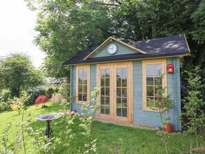 A small, light blue wooden structure with a clock on the front, surrounded by lush greenery and a garden. - Peace Cottage (South Molton)