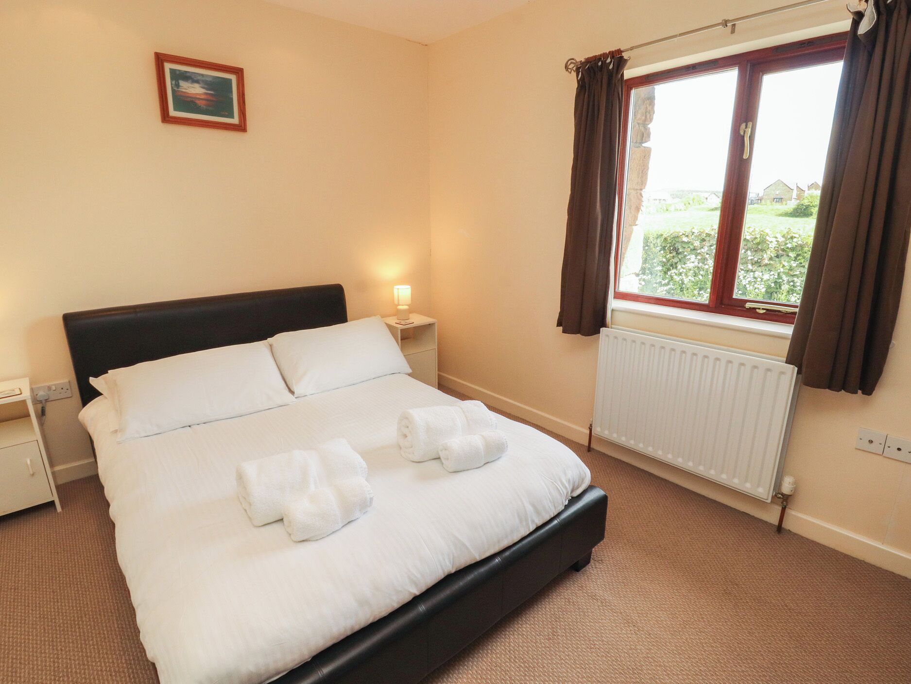 A neatly made bed with white linens and towels, a bedside lamp, a window with curtains, and a radiator.