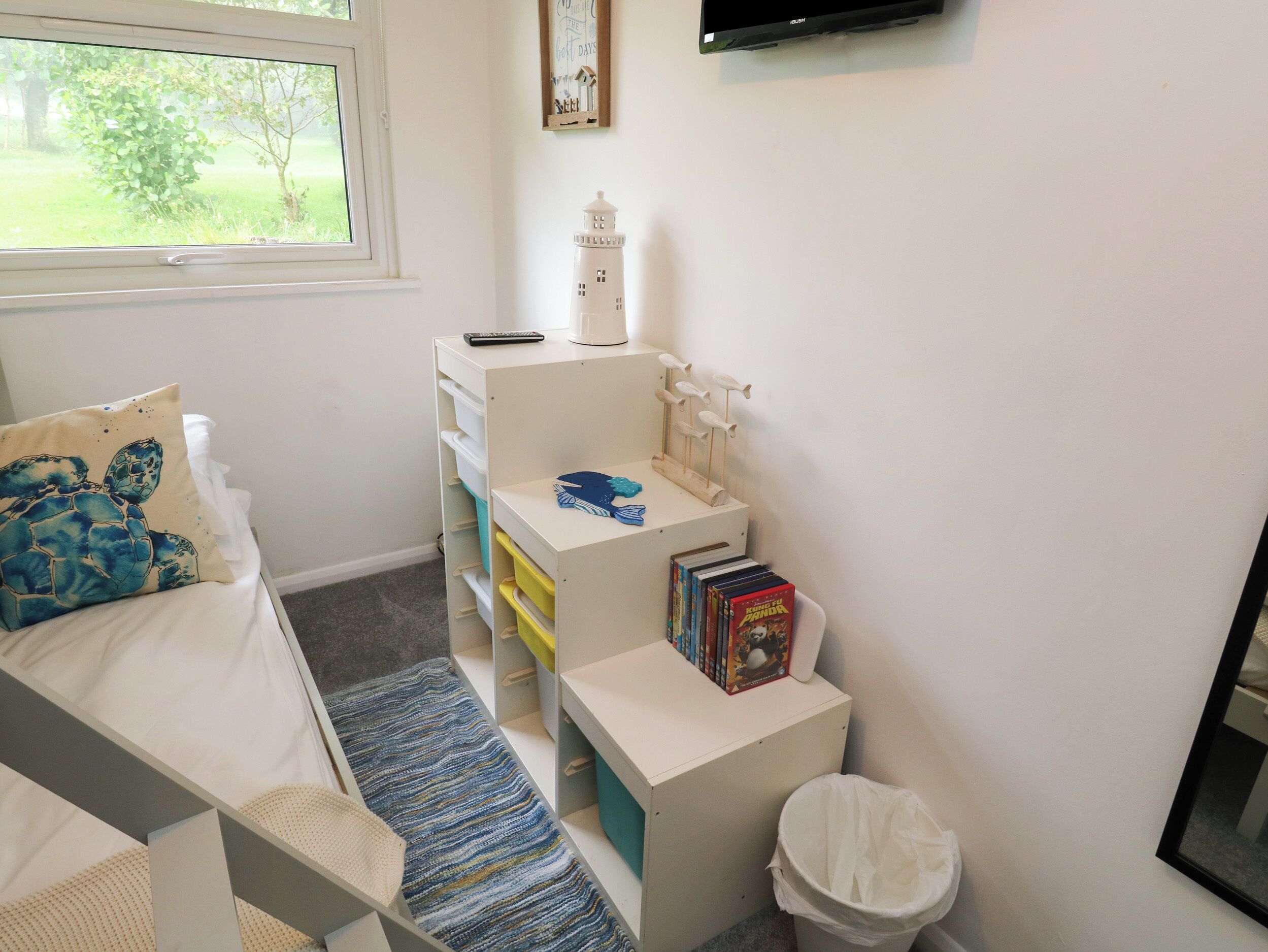 A child's bedroom with a bed, a desk with storage drawers, a bookshelf, a small white trash bin, and a window with a view of greenery.