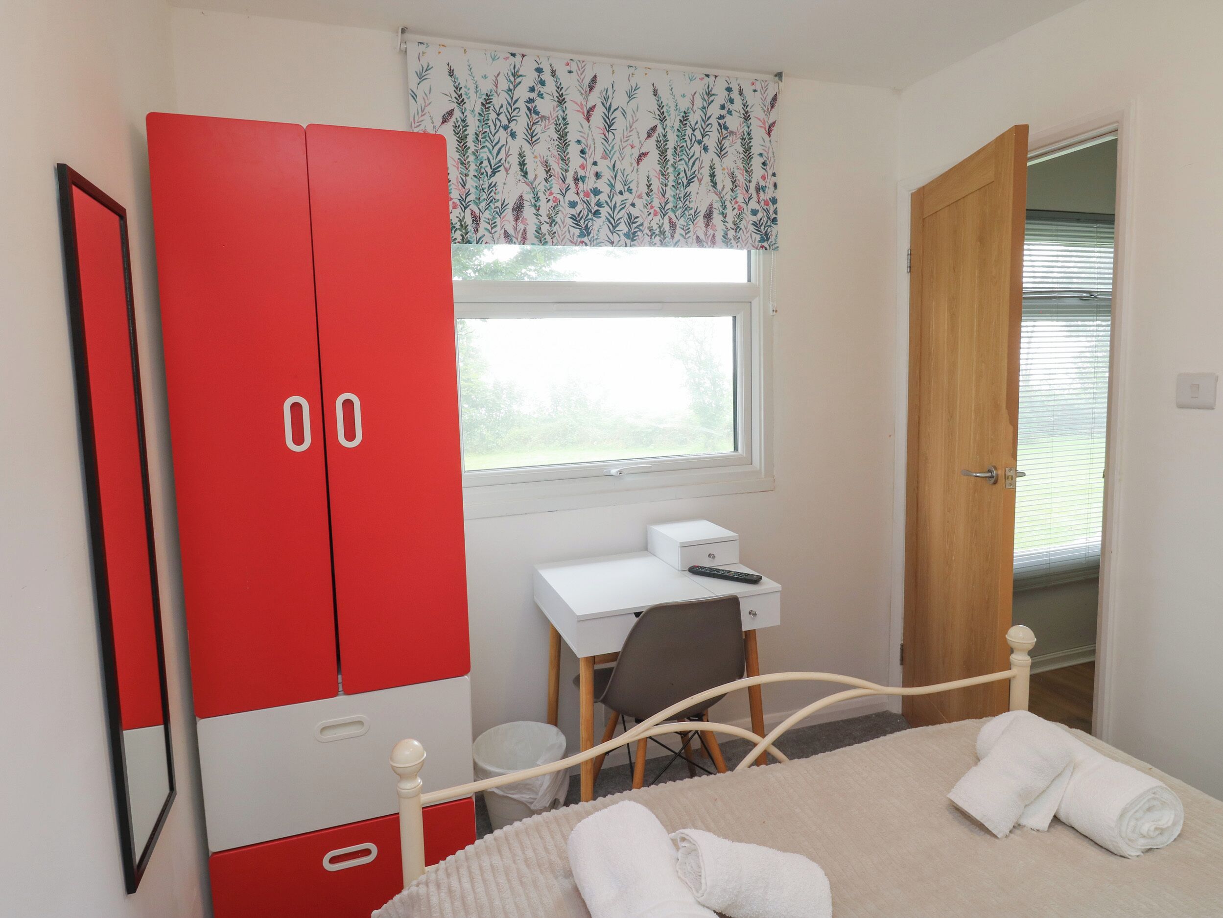 A bedroom with a bed, a desk, a red wardrobe, and a window with a floral curtain.