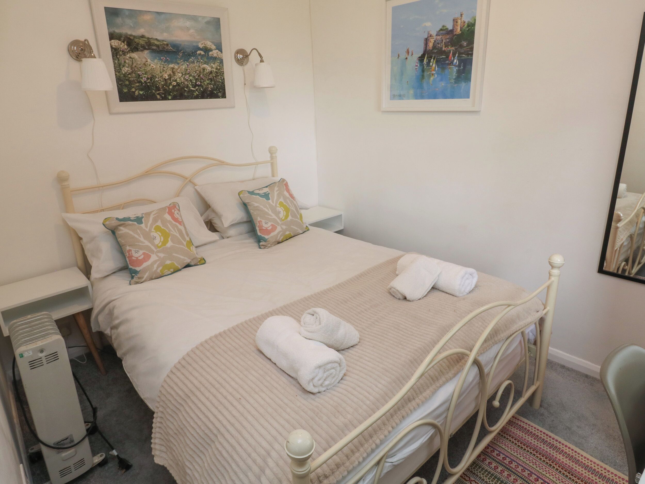 Cama bem arrumada com edredom branco e travesseiros, mesa de cabeceira com abajur e dois quadros emoldurados na parede.