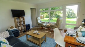 A living room with a black leather sofa, a wooden coffee table, a bookshelf, a television, and a large window with a view of a garden. - 61 Norton Park (Dartmouth)