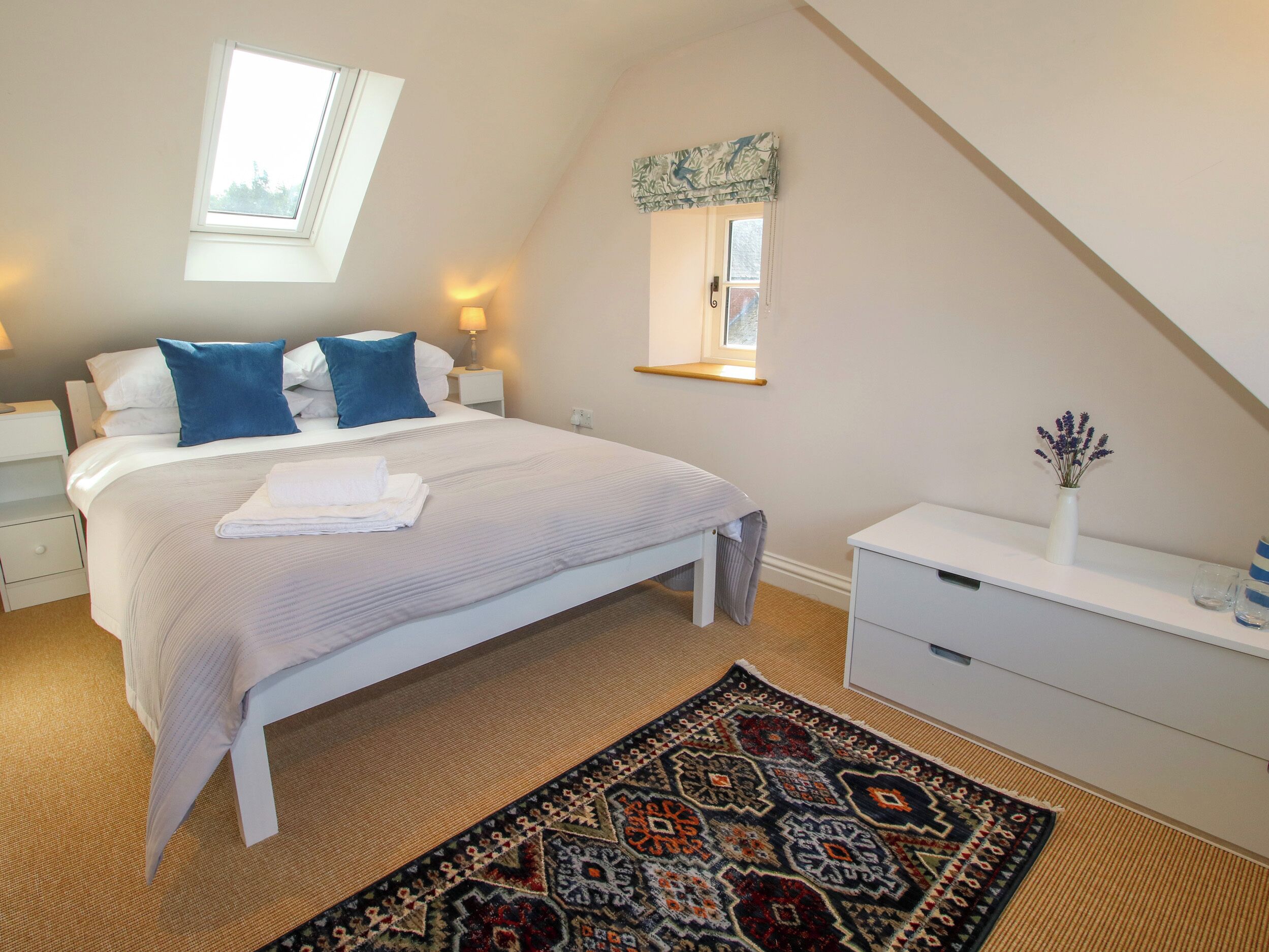 A bedroom with a large bed, a skylight, a bedside table with a lamp, a small table with a vase, and a patterned rug.