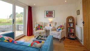 A living room with a blue sofa, striped armchairs, a coffee table with a bottle and glasses, a bookshelf, and a large window with a red curtain. - Rose Cottage (Hereford)