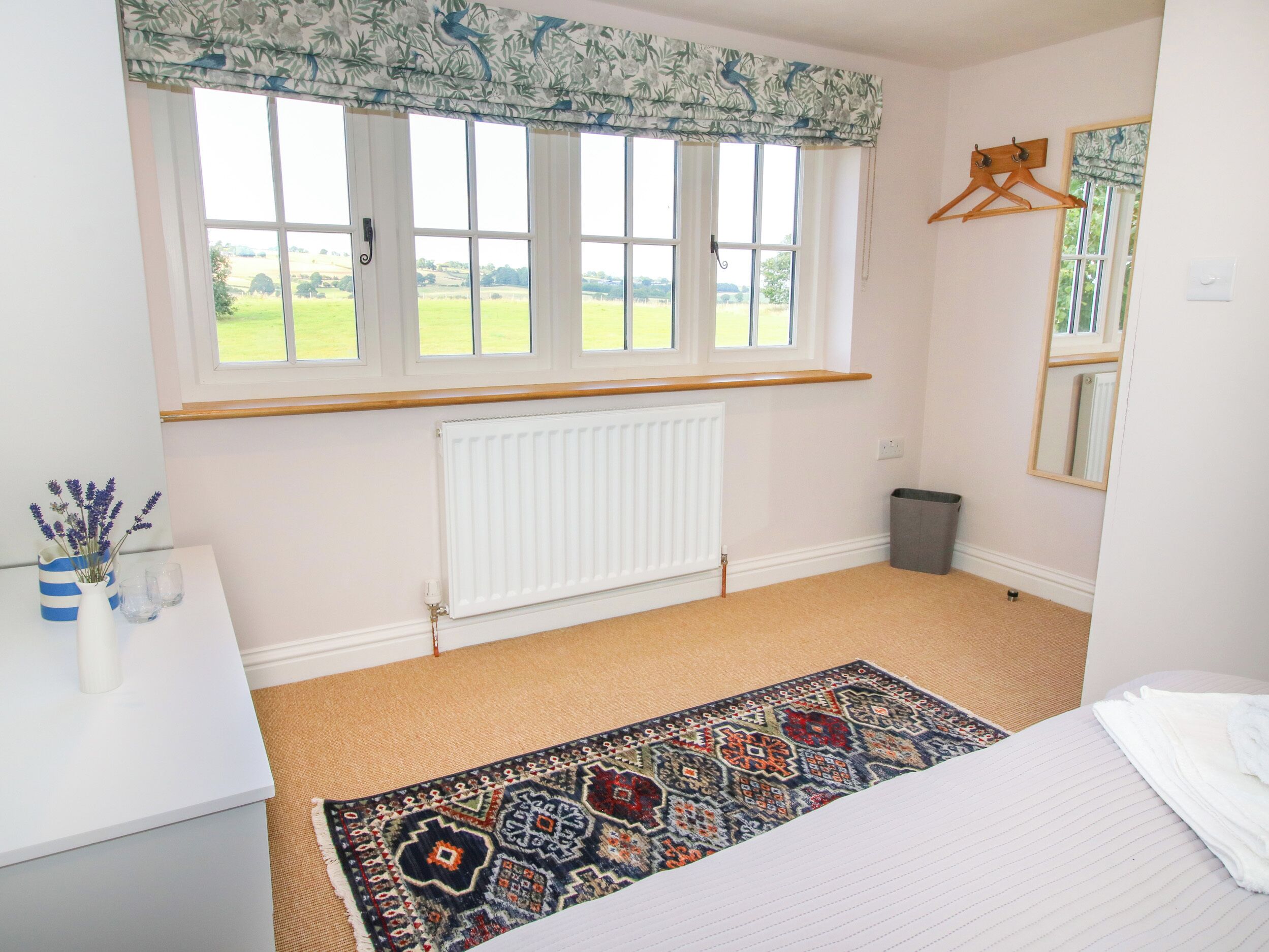 A bedroom with a large window, a radiator, a patterned rug, a bed, and a small table with a vase and glasses.