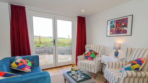 A living room with a blue sofa, striped armchairs, a coffee table with a bottle and glasses, and a view through glass doors to a garden. - Rose Cottage (Hereford)