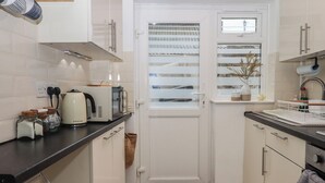 A compact kitchen with white cabinets, a black countertop, a kettle, a toaster, and a window with blinds. - Pie Cottage (Teignmouth)