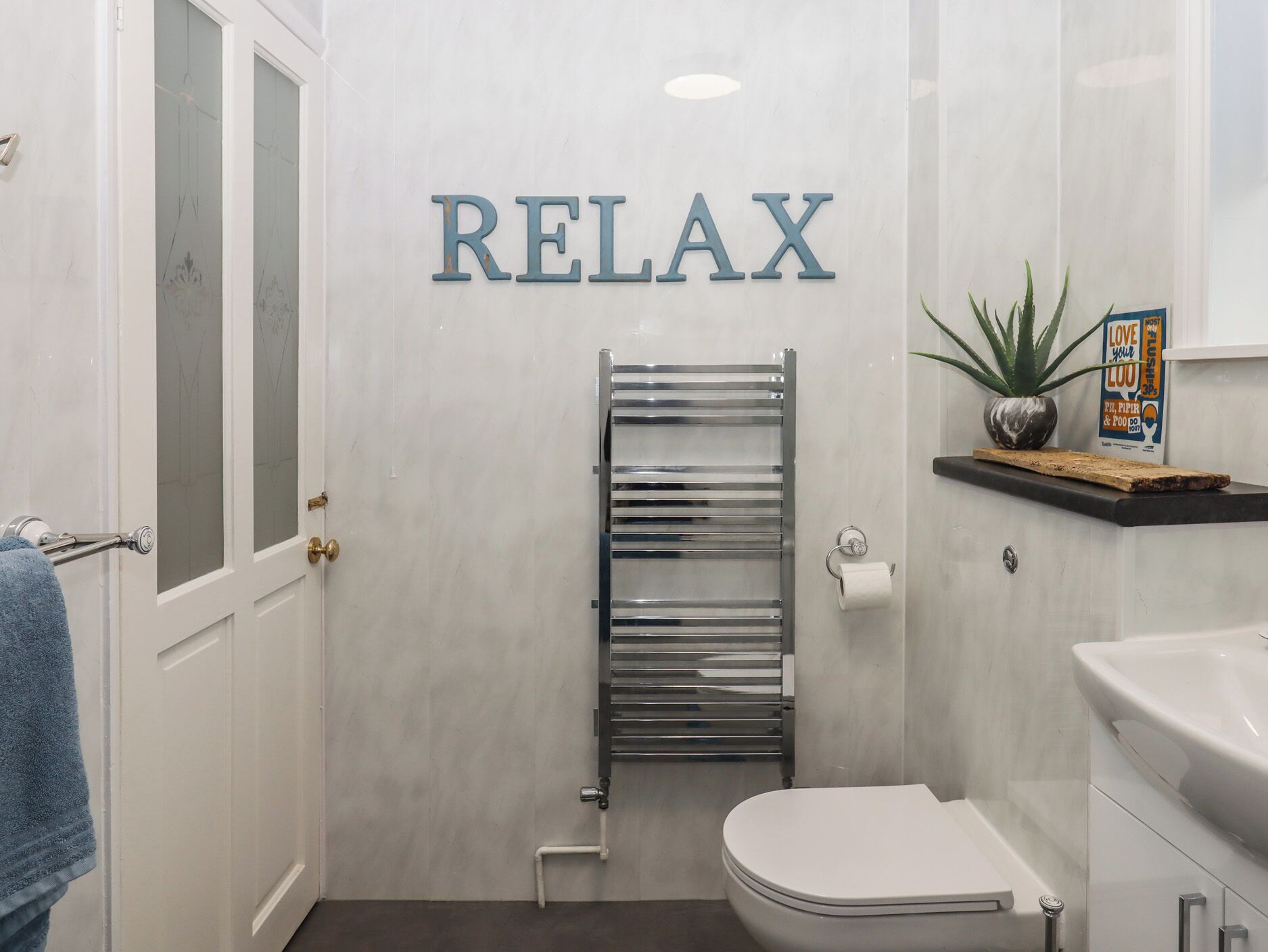 A bathroom with a white toilet, a towel rack, a towel, a shower with a glass door, and a sign that says "RELAX".