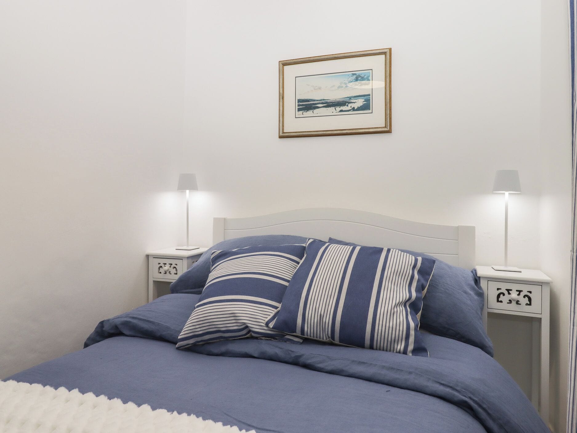 A neatly made bed with blue bedding and striped pillows, two bedside tables with lamps, and a framed picture on the wall.