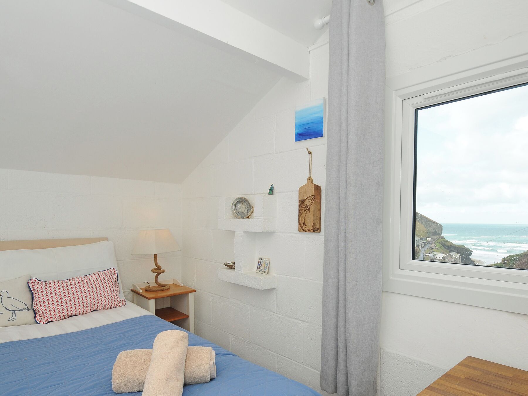 Ein Schlafzimmer mit Bett, Nachttisch, Lampe, Fenster mit Meerblick und Wanddekorationen.