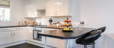 A modern kitchen with white cabinets, a black countertop, and a stainless steel range hood. There is a black bar stool and a fruit display on the counter.