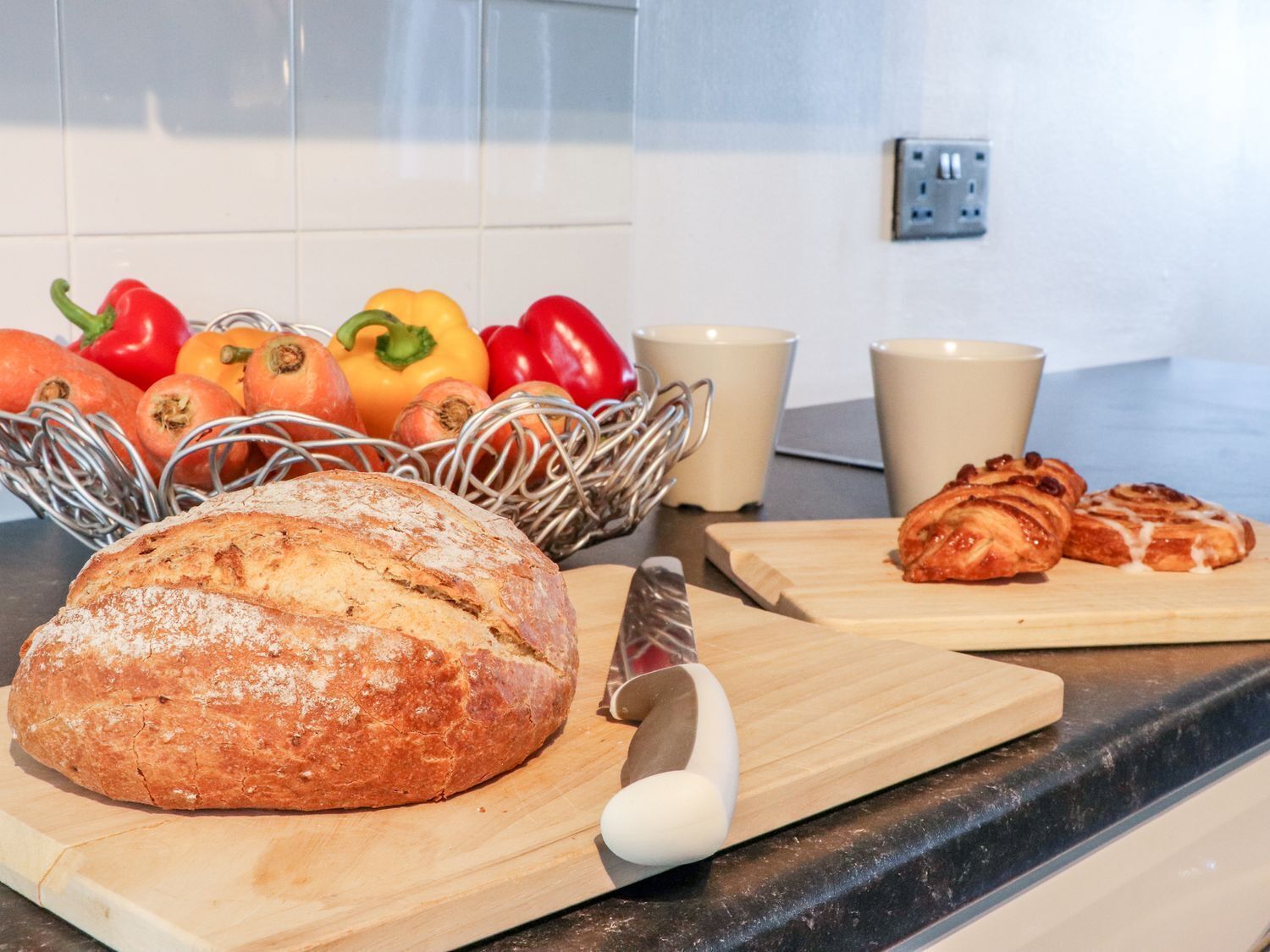 A kitchen counter with a loaf of bread, a basket of vegetables, and a cutting board with pastries.