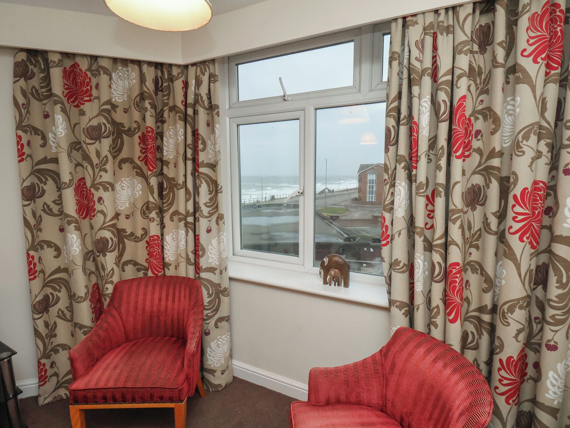 A room with two red armchairs, a window with a view of the sea, and floral curtains.