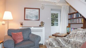Ein gemütliches Wohnzimmer mit einer grauen Couch, einem kuscheligen Ohrensessel, einem Bücherregal, einem gerahmten Bild und einem Fenster mit Blick auf eine felsige Landschaft.