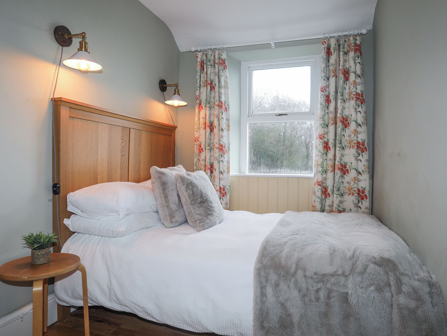 A bedroom with a wooden headboard, a bed with white bedding, two fluffy pillows, a bedside table with a plant, and a window with floral curtains.