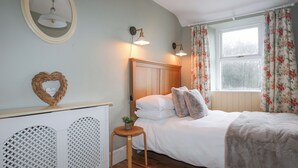 Ein Schlafzimmer mit einem Holzkopfteil, einem Bett mit weißer Bettwäsche, einem kleinen runden Tisch mit einer Pflanze, einer herzförmigen Dekoration und einem Fenster mit floralen Vorhängen.