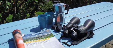 A blue table with a magazine, two blue cups, a coffee maker, and binoculars.