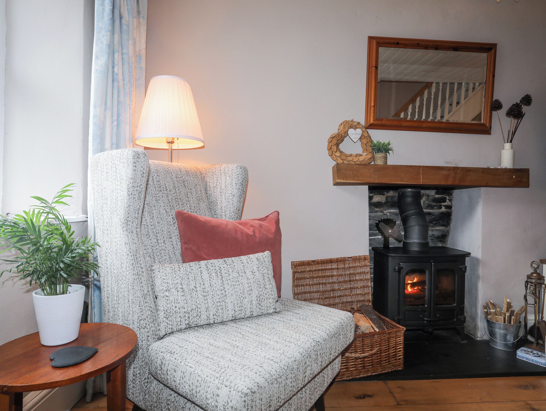 Un salón acogedor con un sillón estampado, una mesita con planta en maceta, una chimenea con manto de piedra y un espejo en la pared.