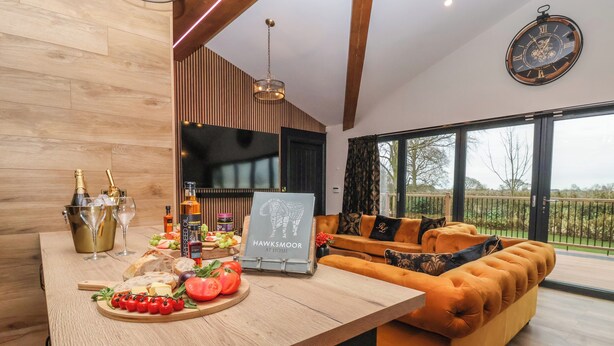 A modern living room with a wooden floor, a sofa, a dining area with a wooden table, and a large window offering a view of the outdoors. - Routhorpe Lodge (Driffield)