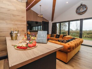 A modern living room with a wooden floor, a sofa, a dining area with a wooden table, and a large window offering a view of the outdoors. - Routhorpe Lodge (Driffield)