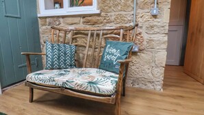 Une chaise en bois rustique, agrémentée d’un coussin à motifs floraux et de deux coussins décoratifs, est placée contre un mur de pierre, juste en dessous d’une fenêtre.