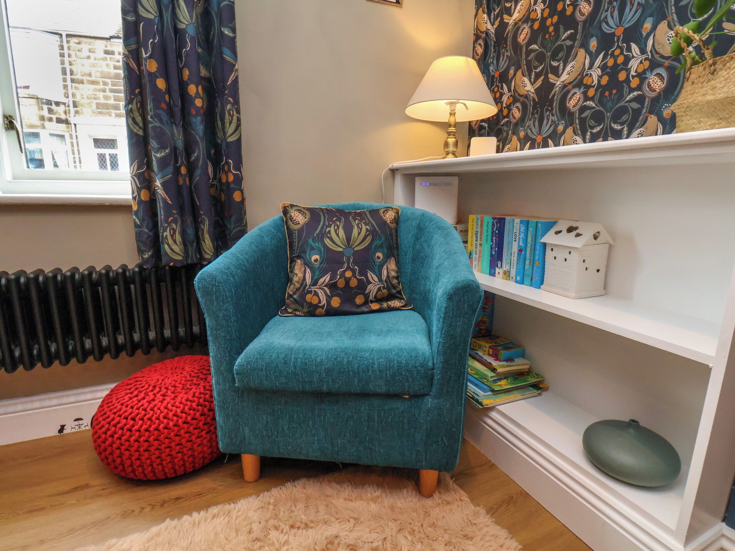 Uma poltrona azul-esverdeada com uma almofada decorativa, um banquinho vermelho de tricô, um tapete branco e fofo, uma estante com livros e uma pequena maquete de casa, e um radiador.