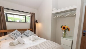 Un lit bien fait, avec des oreillers et des serviettes, une lampe de chevet, une fenêtre avec des rideaux, une petite table sur laquelle est posé un vase de tournesols, et une armoire avec des cintres.