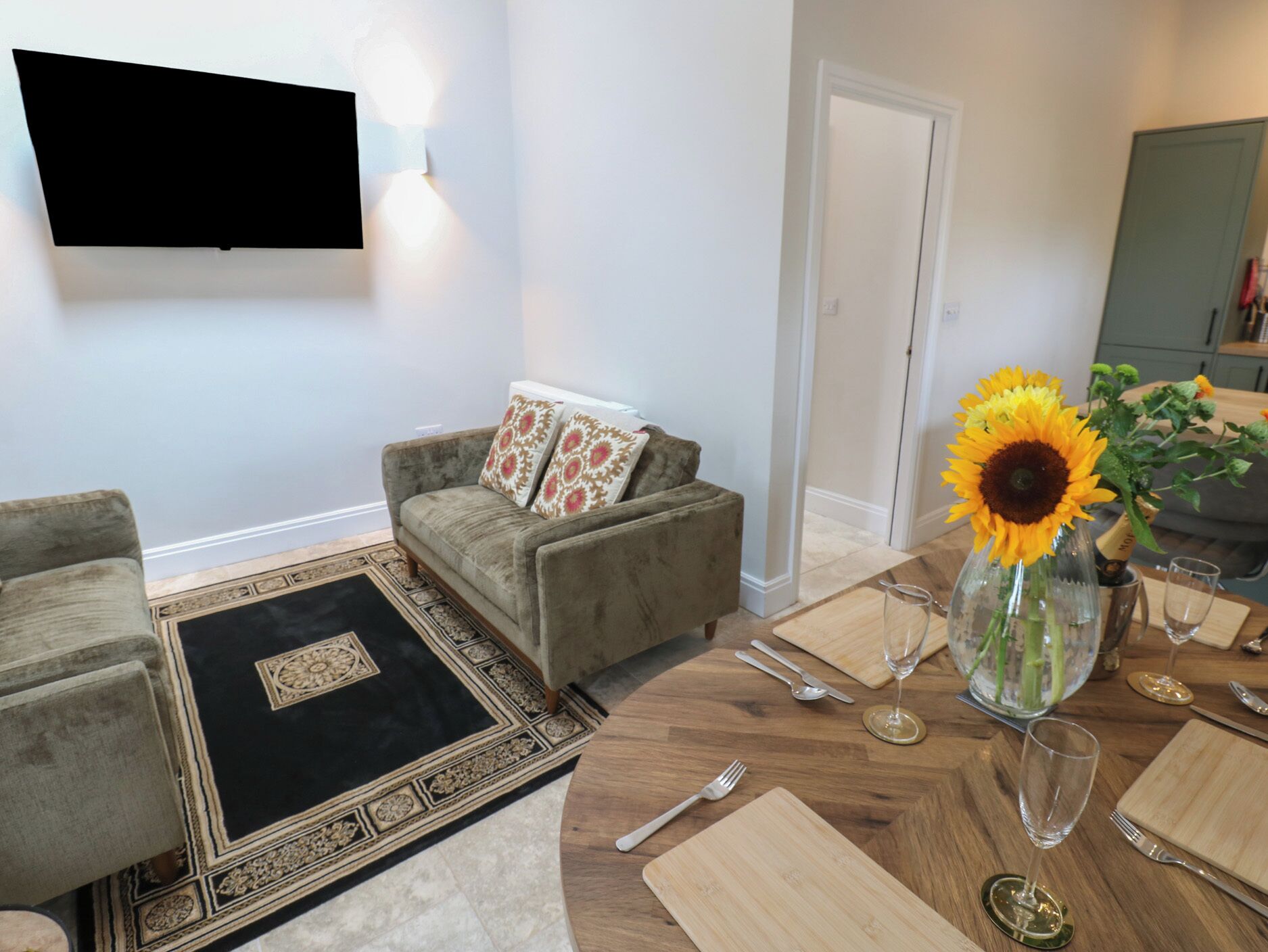A living room with a sofa, a coffee table, and a dining table set for two with cutlery and a vase with a sunflower.