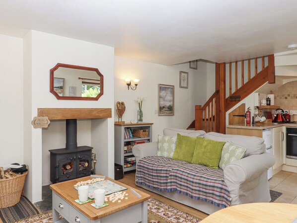 A cozy living room with a fireplace, a sofa with green cushions, a coffee table with a board game, and a kitchen area with a wooden staircase. - Dove Cottage (South Molton)