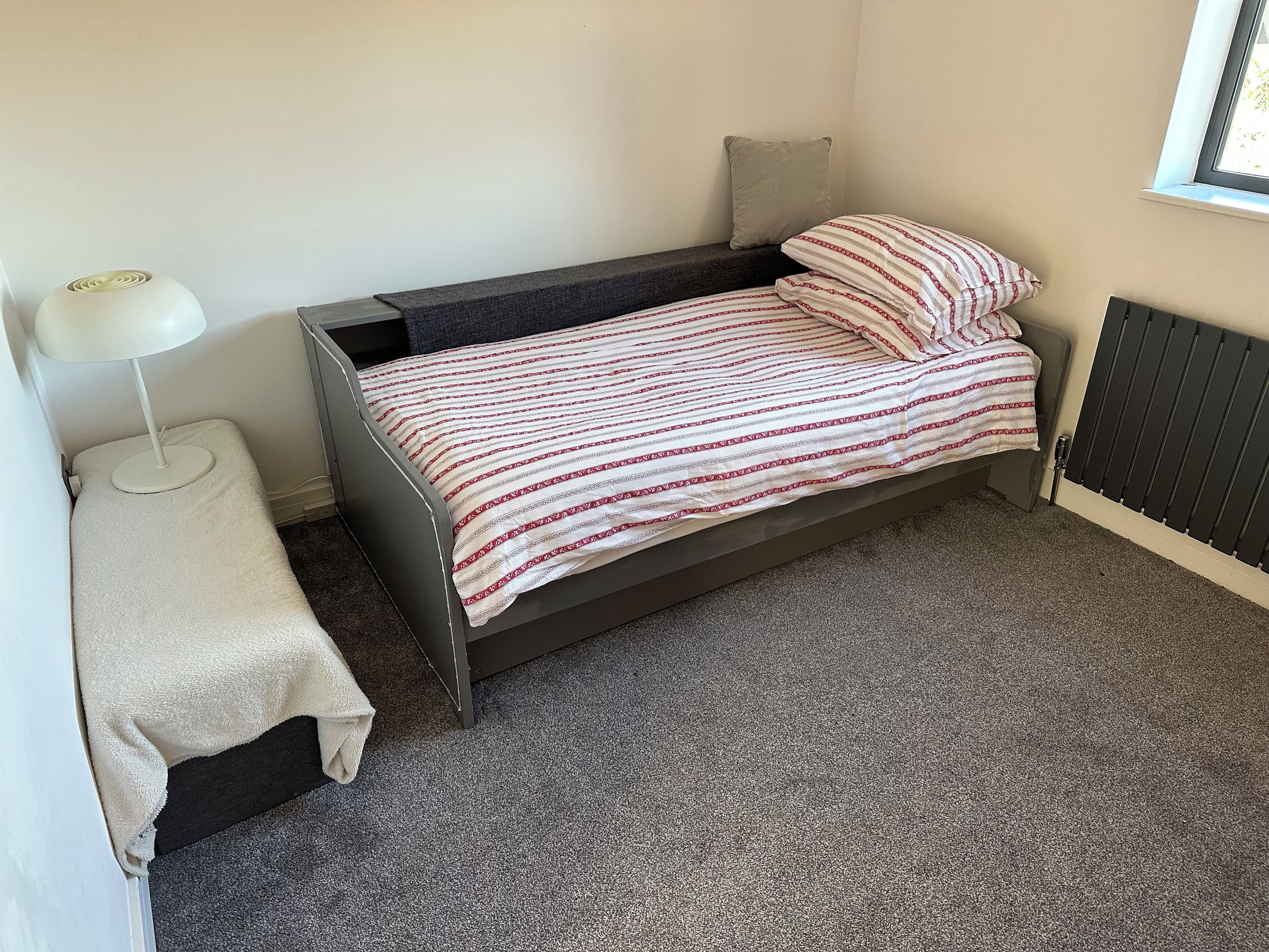 A small, single-bed room with a grey carpet, a white bedside table, a white lamp, and a window with blinds.