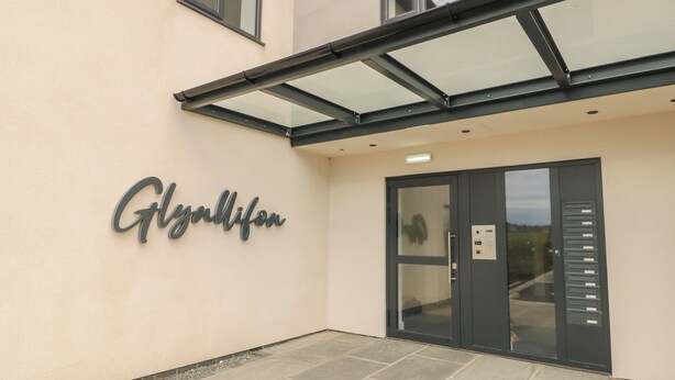 Entrada de un edificio con toldo de vidrio, una placa con el nombre "Glynnifon" y una puerta con panel de control.