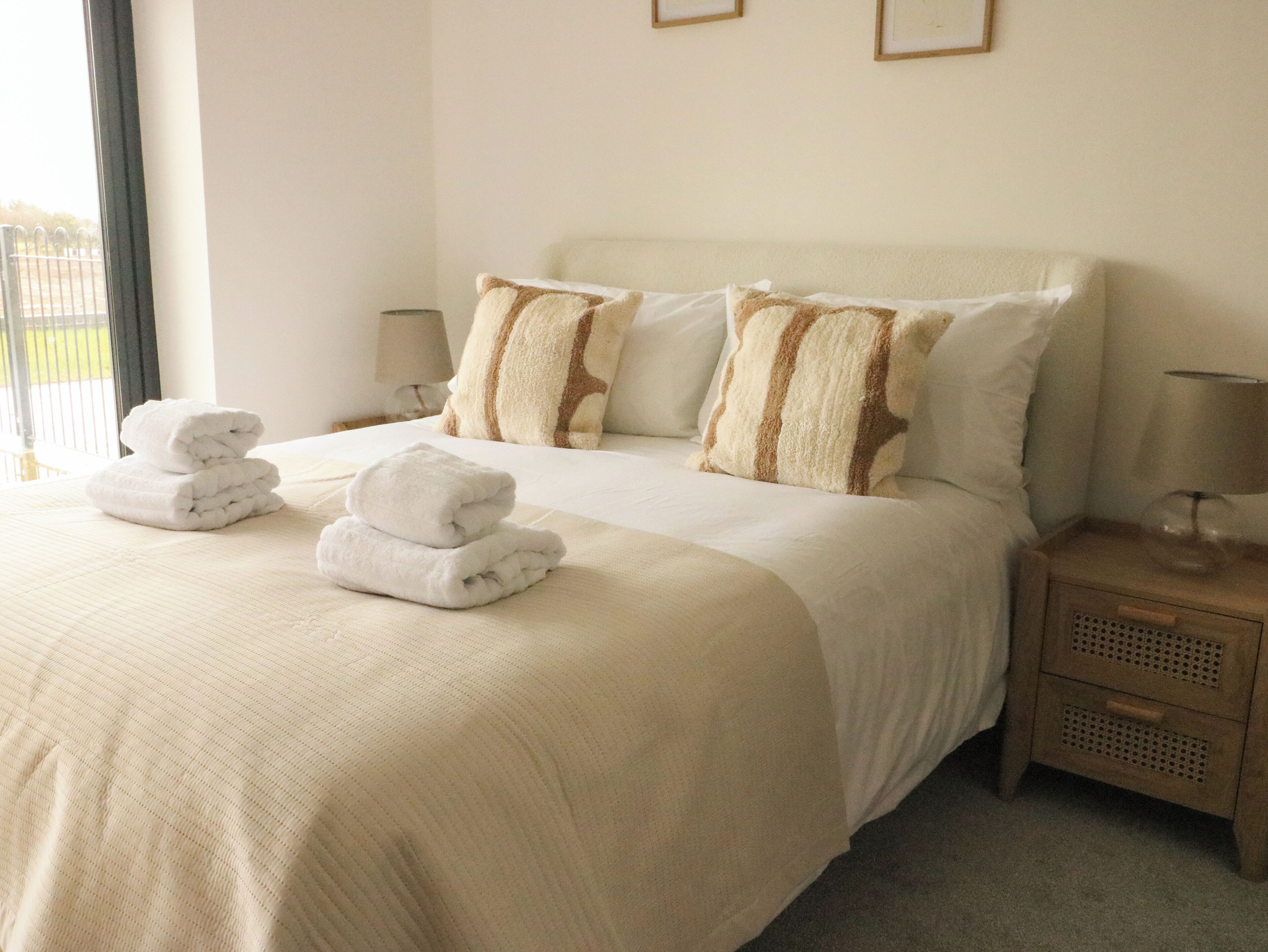 A neatly made bed with white linens and two pillows, a bedside table with a lamp, and a view of the outdoors through a window.