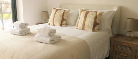 A neatly made bed with white linens and two pillows, a bedside table with a lamp, and a view of the outdoors through a window.