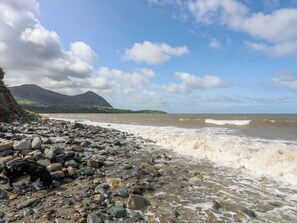 Ein felsiger Strand mit brechenden Wellen, ein Berg im Hintergrund und ein teils bewölkter Himmel.