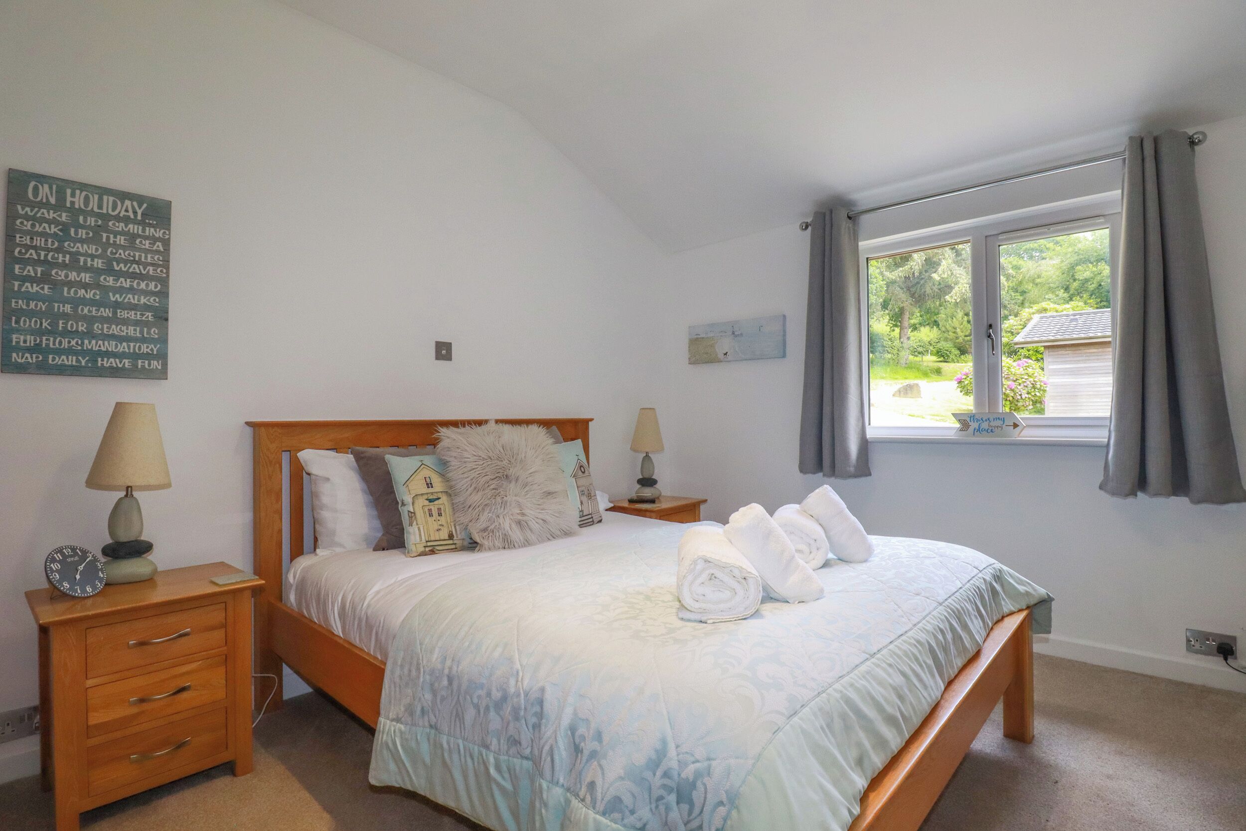 Un dormitorio con cama, dos mesitas de noche, una ventana con cortinas, un reloj de pared y un letrero con consejos para las vacaciones.