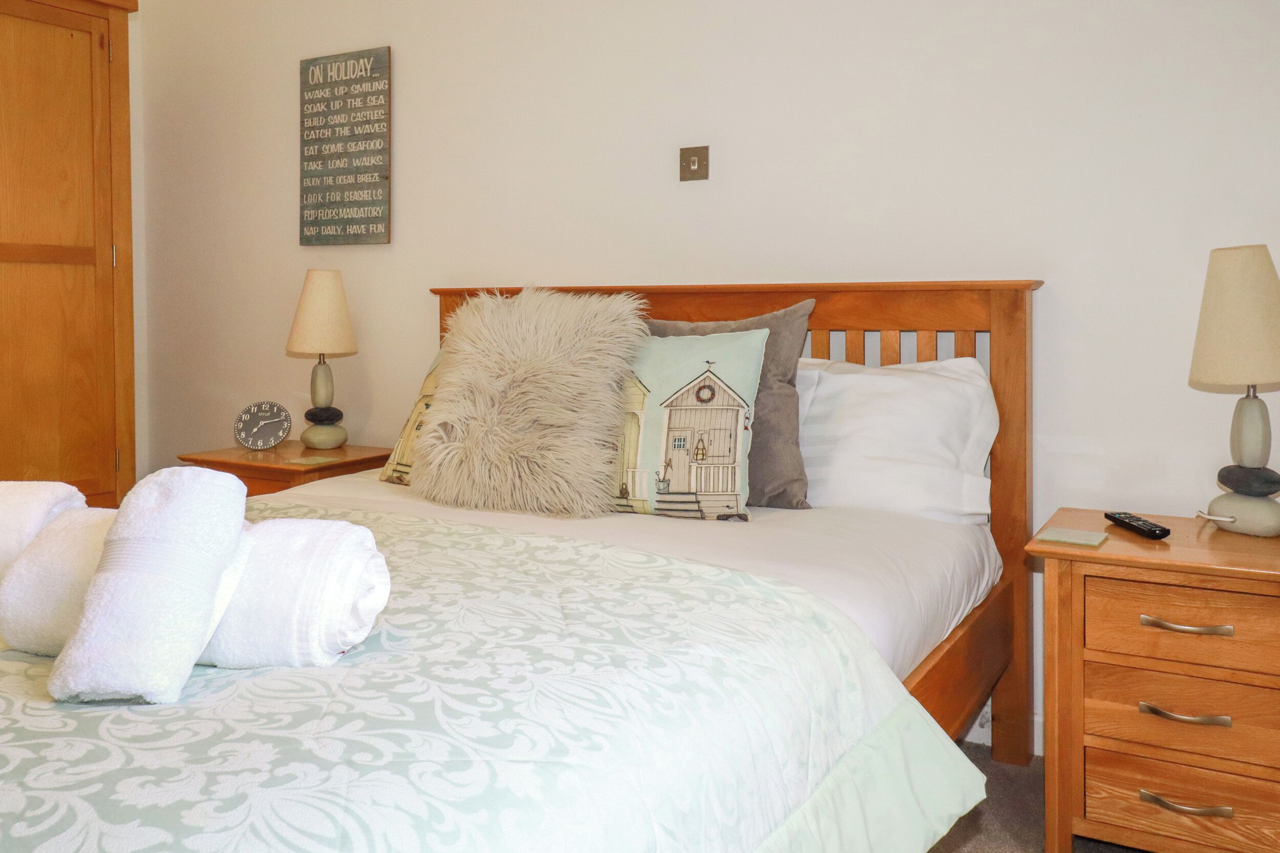 A neatly made bed with a patterned bedspread, two pillows, a fluffy throw, and a bedside lamp. There is a small table with a clock and a decorative item beside the bed.