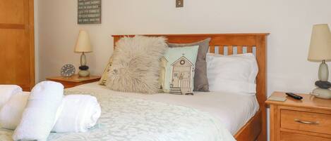 A neatly made bed with a patterned bedspread, two pillows, a fluffy throw, and a bedside lamp. There is a small table with a clock and a decorative item beside the bed.