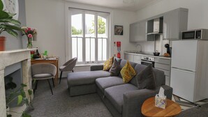 Ein modernes Wohnzimmer mit einem grauen Sofa, einem kleinen Holztisch und einer Küchenzeile mit weißen Geräten.