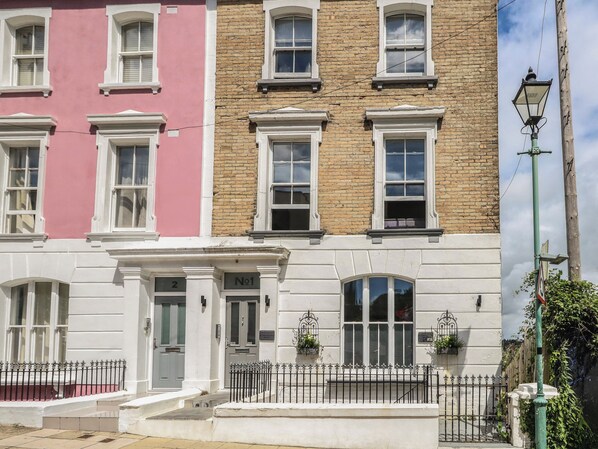 Une rangée de maisons de ville aux façades blanches et roses, avec des portes numérotées et un petit jardin devant, délimité par une clôture en métal.