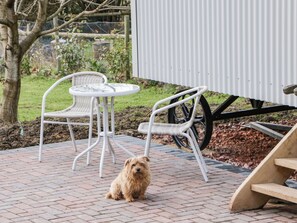 Un patio con una mesa redonda pequeña y dos sillas blancas, un perro sentado en el suelo, y un banco de madera.