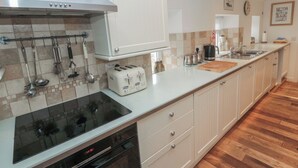 A modern kitchen with a stainless steel range hood, a built-in oven, and a dishwasher. There is a white countertop with a toaster and various kitchen utensils hanging above it. The kitchen has white cabinets and a tiled backsplash. - The Smithy (Chathill)