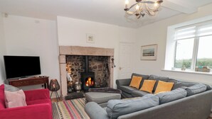 A living room with a stone fireplace, a grey sofa, a red armchair, and a television on a wooden cabinet. - The Smithy (Chathill)