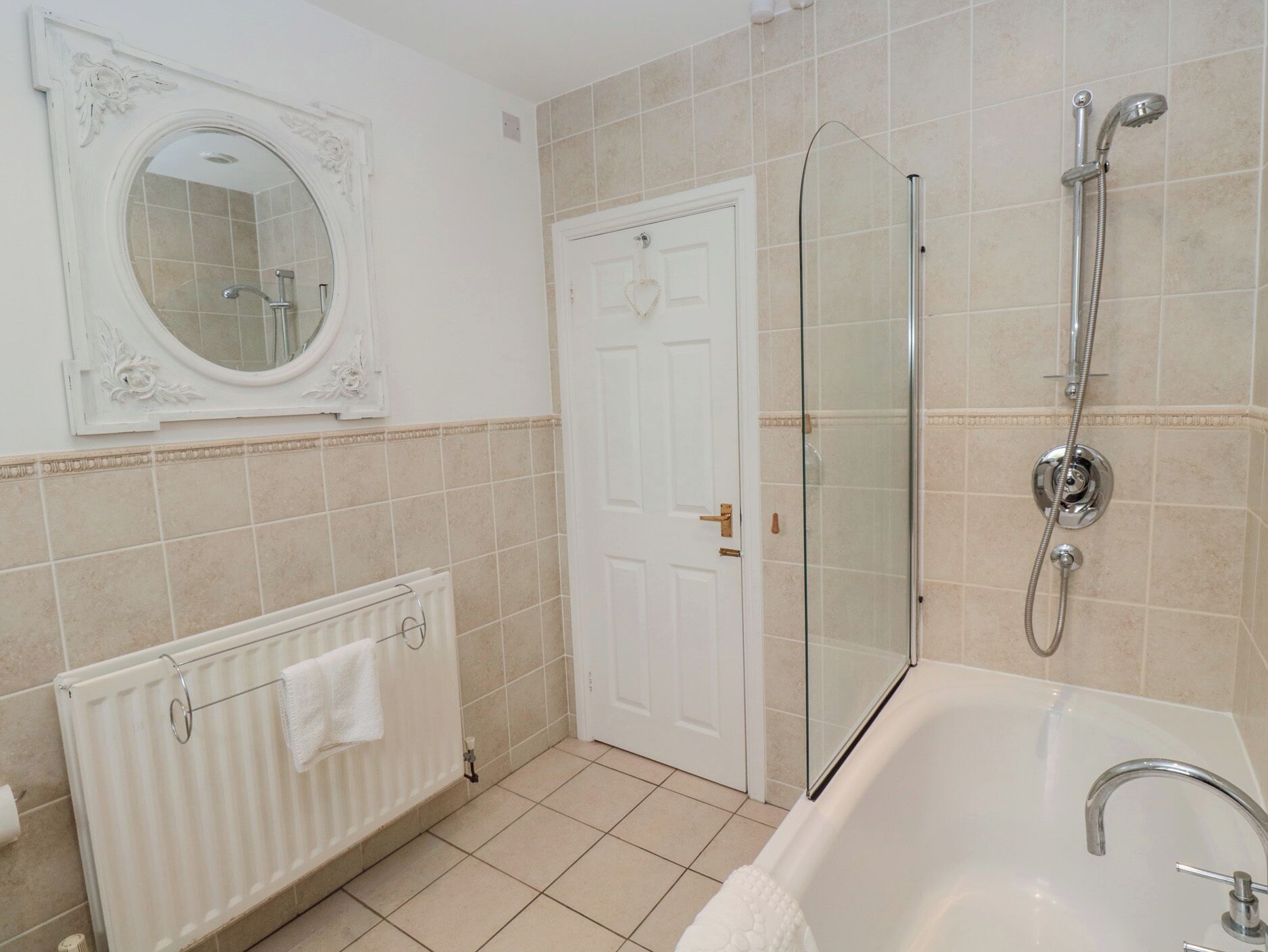 Een badkamer met een badkuip, een douche met glazen wand, een witte deur en een ronde spiegel met decoratieve details.