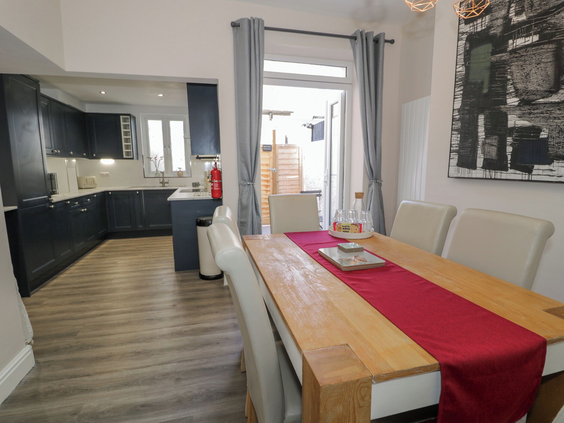 Uma cozinha moderna com armários escuros, uma mesa de jantar de madeira com um caminho vermelho e vista para a área de refeições através de uma porta de vidro.
